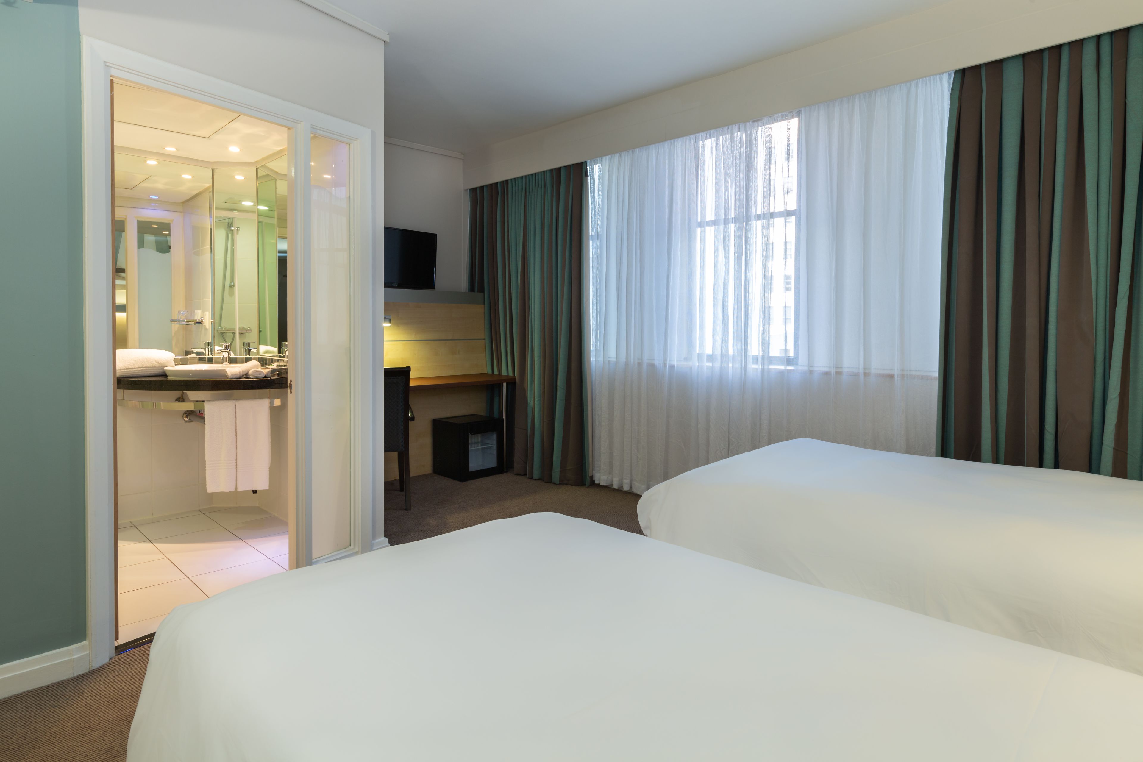A hotel room with two beds, a desk, a TV, and a bathroom with a mirror and sink.