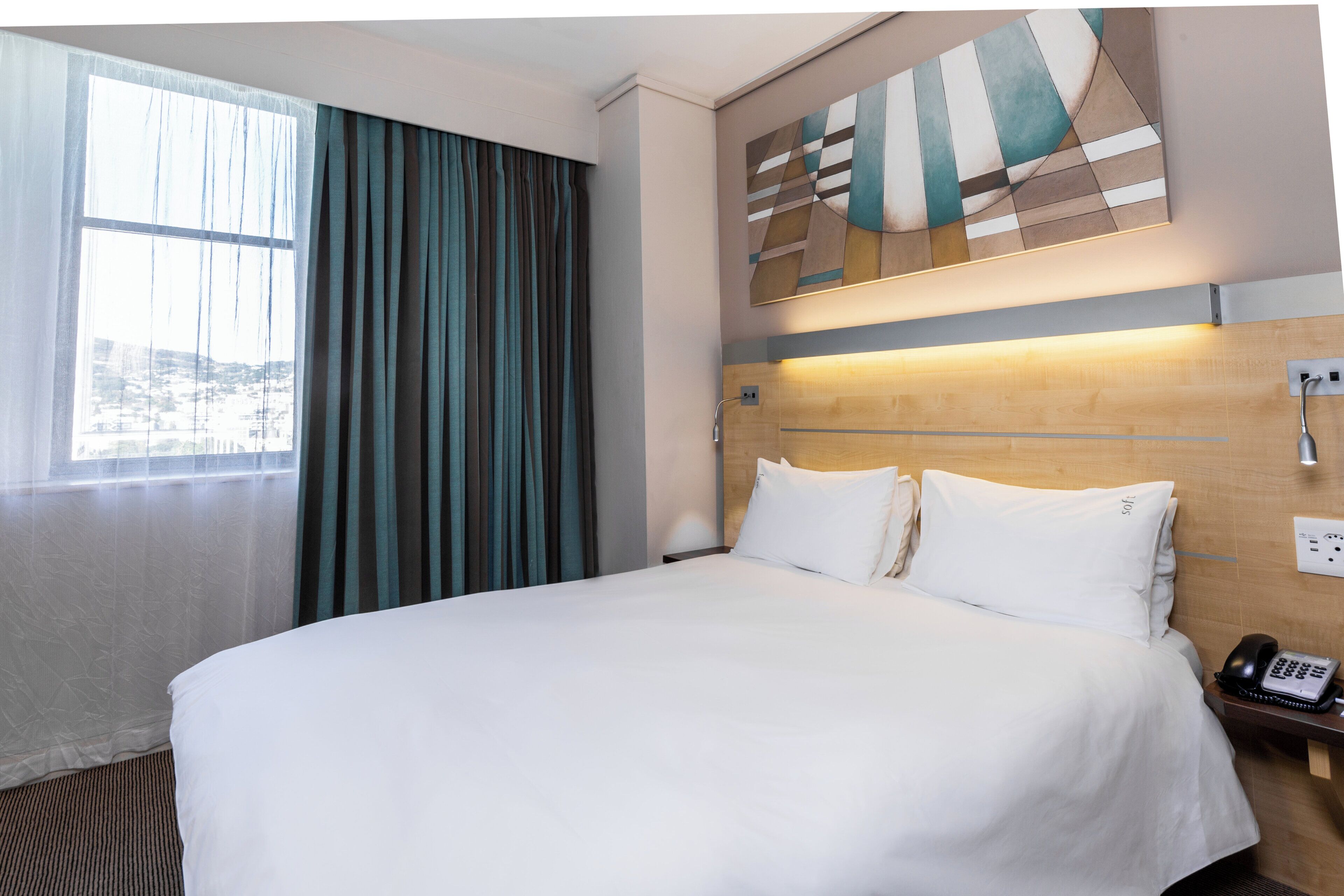 Une chambre d’hôtel avec un grand lit, une vue sur la ville par la fenêtre et une peinture abstraite moderne accrochée au mur.