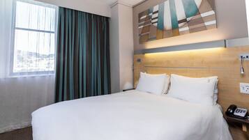 Une chambre d’hôtel avec un grand lit, une vue sur la ville par la fenêtre et une peinture abstraite moderne accrochée au mur.