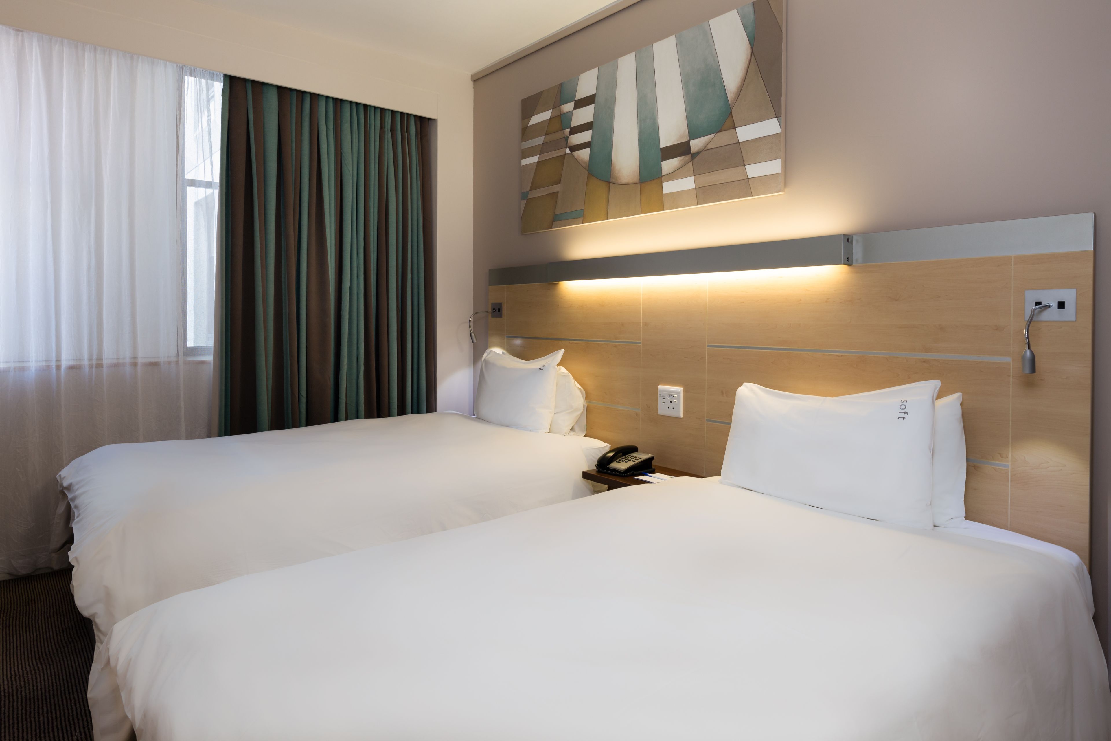 A hotel room with two beds, a large window with curtains, a wall-mounted telephone, and a modern abstract painting.