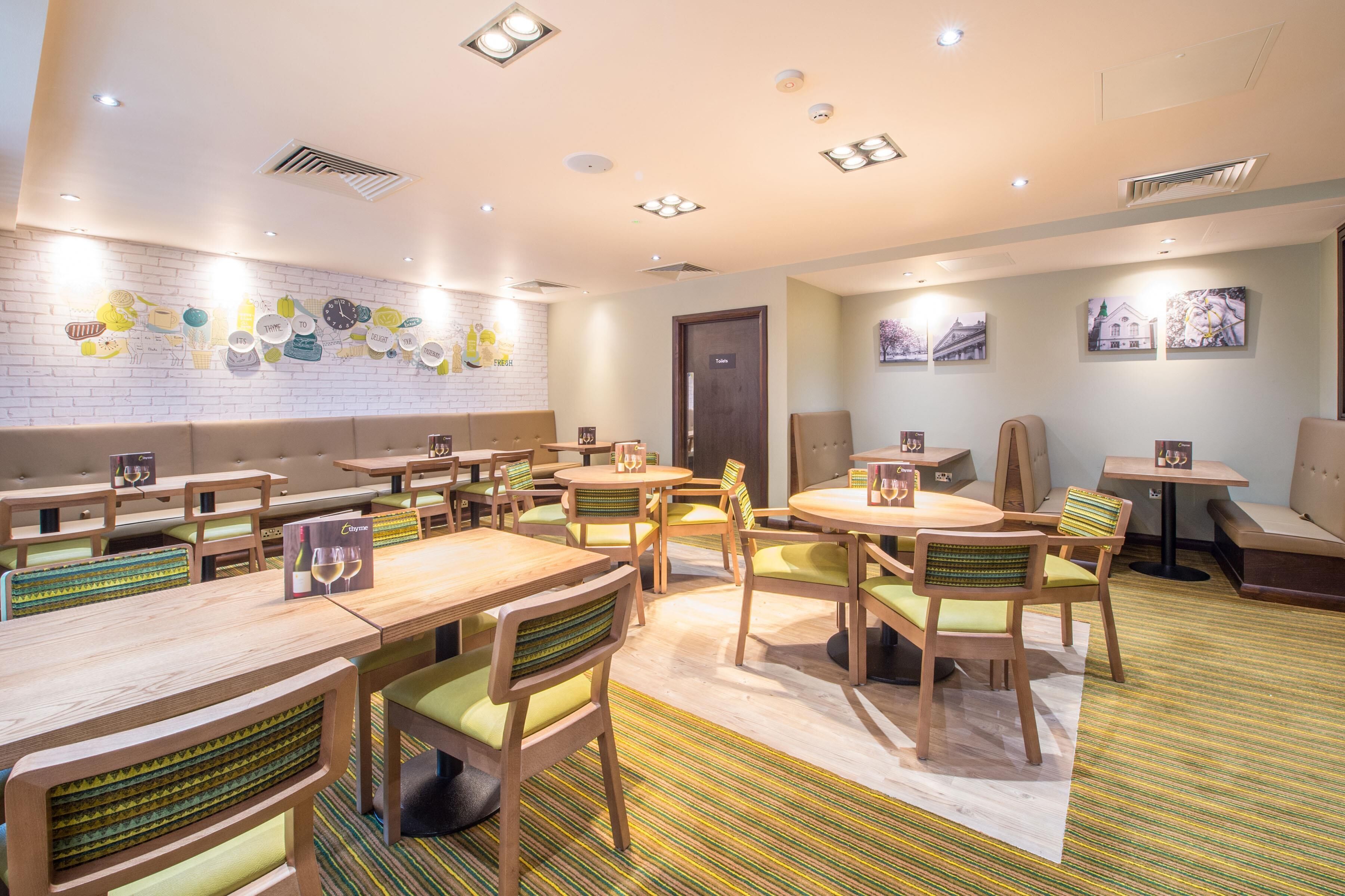 A hotel dining area with multiple tables and chairs, a brick wall with artwork, and a door labeled 'Lobby'.
