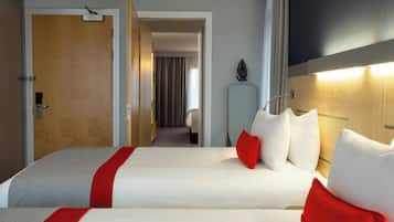 Ein Hotelzimmer mit zwei Betten, einer Holztür, einem weißen Kopfteil und einem Nachttisch mit einer Lampe.