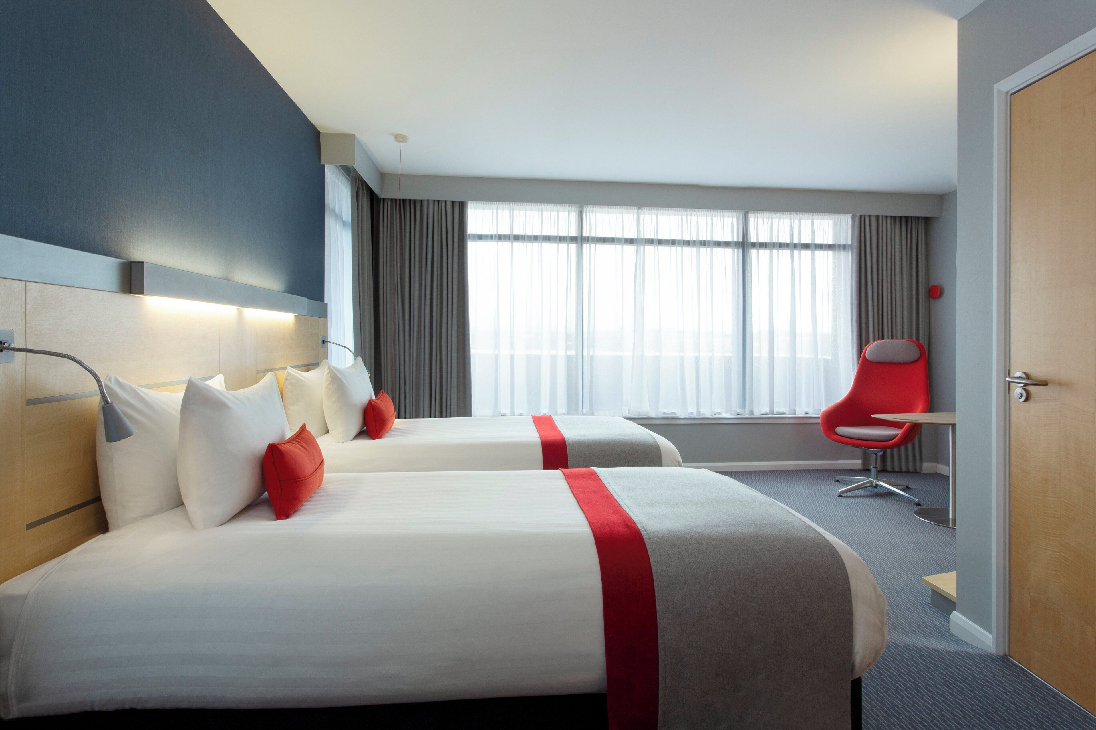 A hotel room with two beds, a red chair, and a large window with curtains.