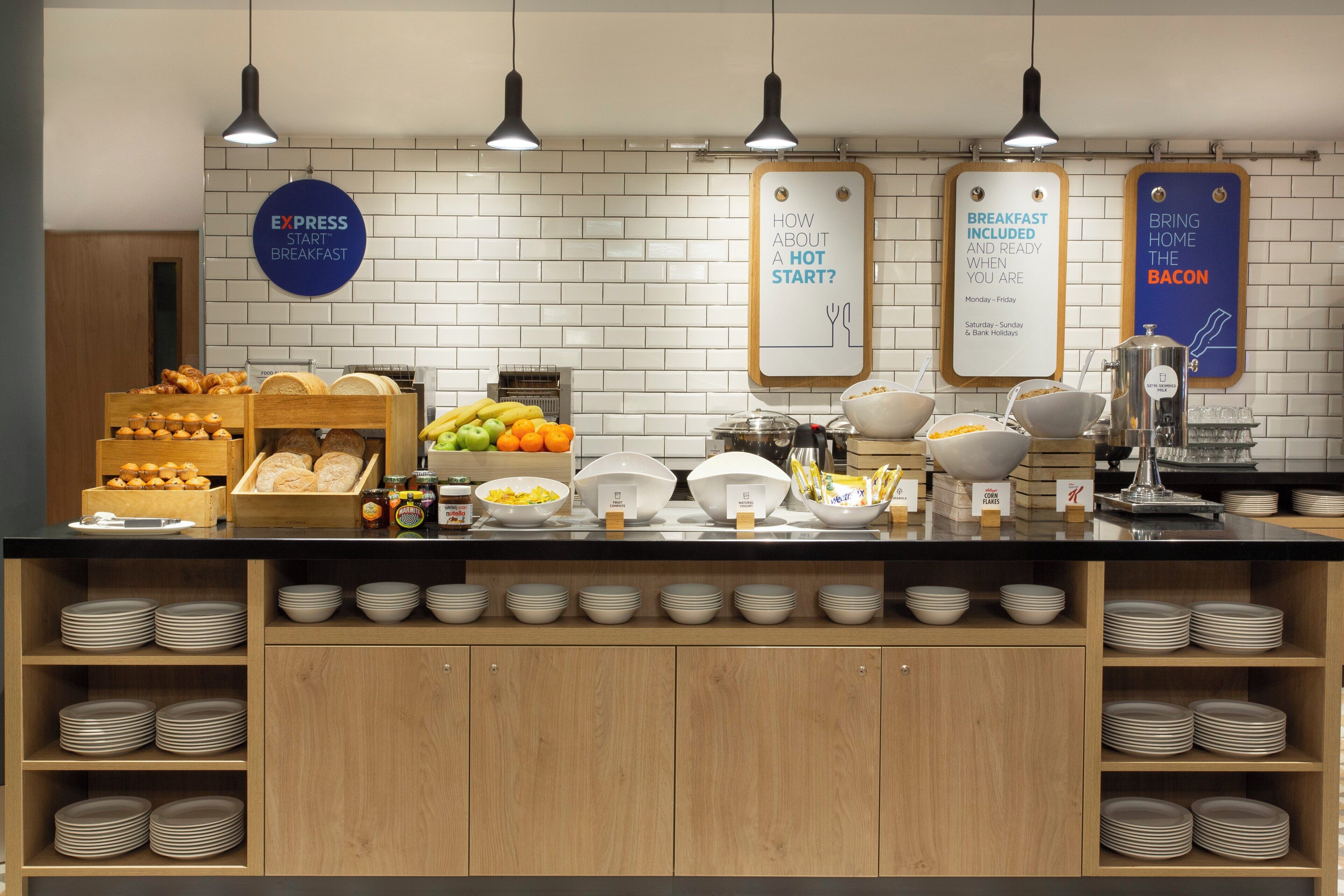 A breakfast buffet with labeled food stations, including bread, fruits, cereals, and dairy products.