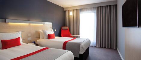 Una habitación de hotel con dos camas, una silla roja, un televisor de pantalla plana y un ventanal con cortinas.