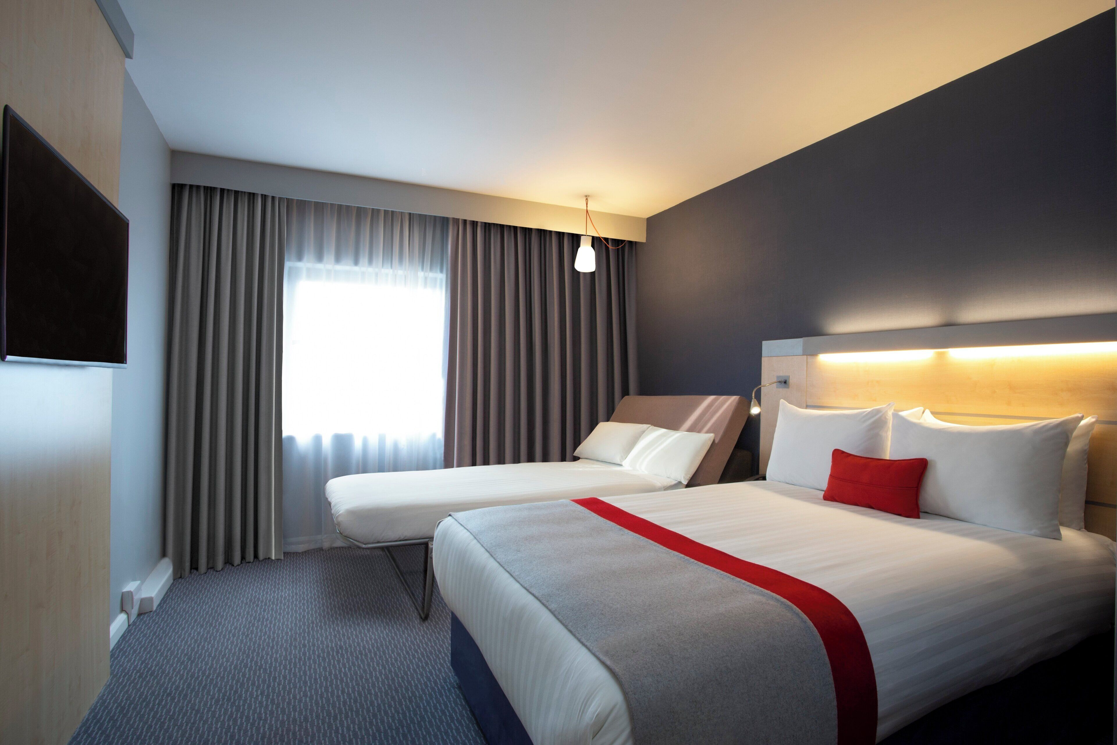 A hotel room with two beds, a large window with curtains, a flat-screen TV, and a modern headboard.