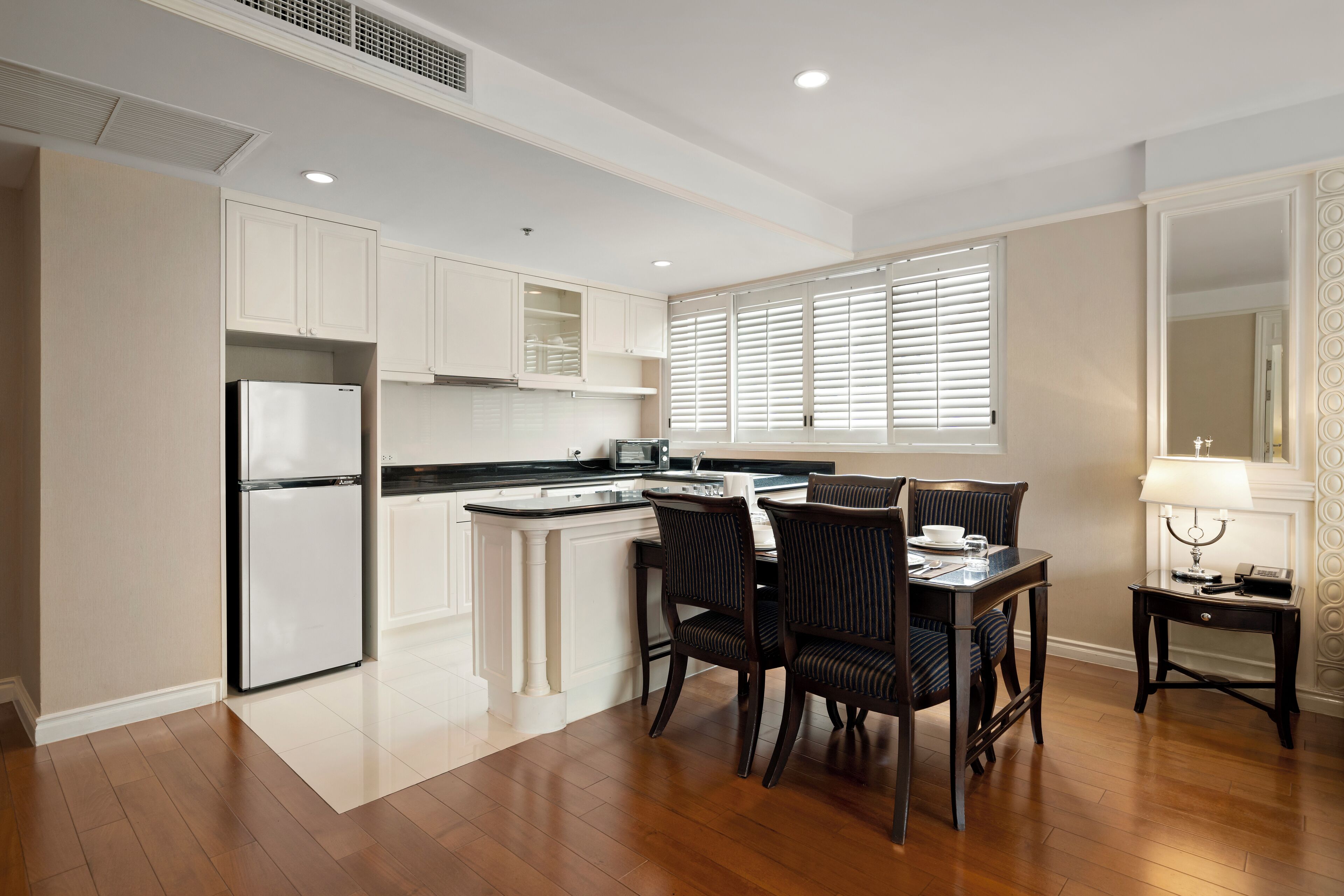 A modern kitchen with white cabinetry, a built-in refrigerator, a dining area with two chairs, and a small table with a lamp.