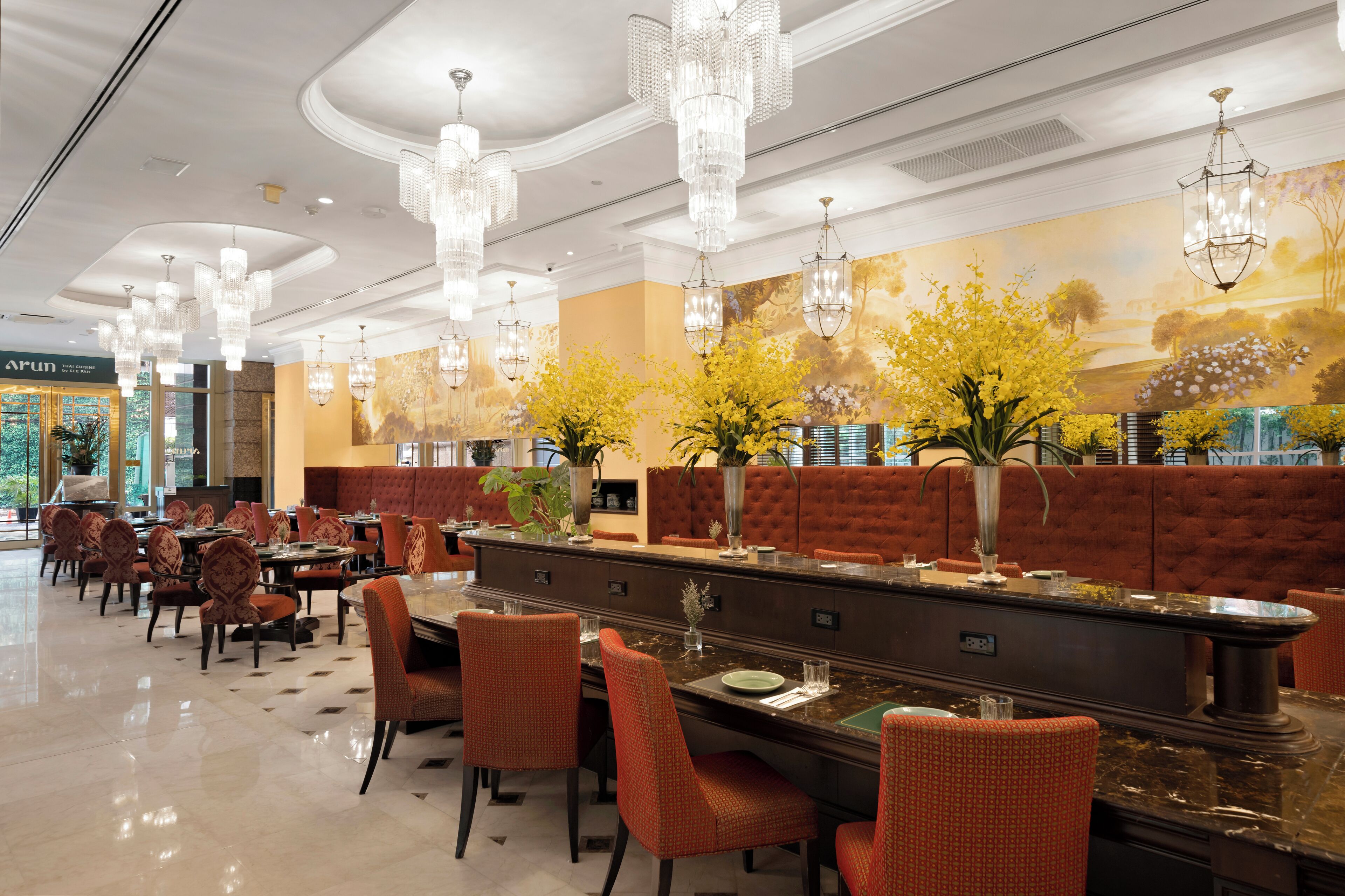 A spacious restaurant with high ceilings, ornate chandeliers, and a long counter with red upholstered seating.
