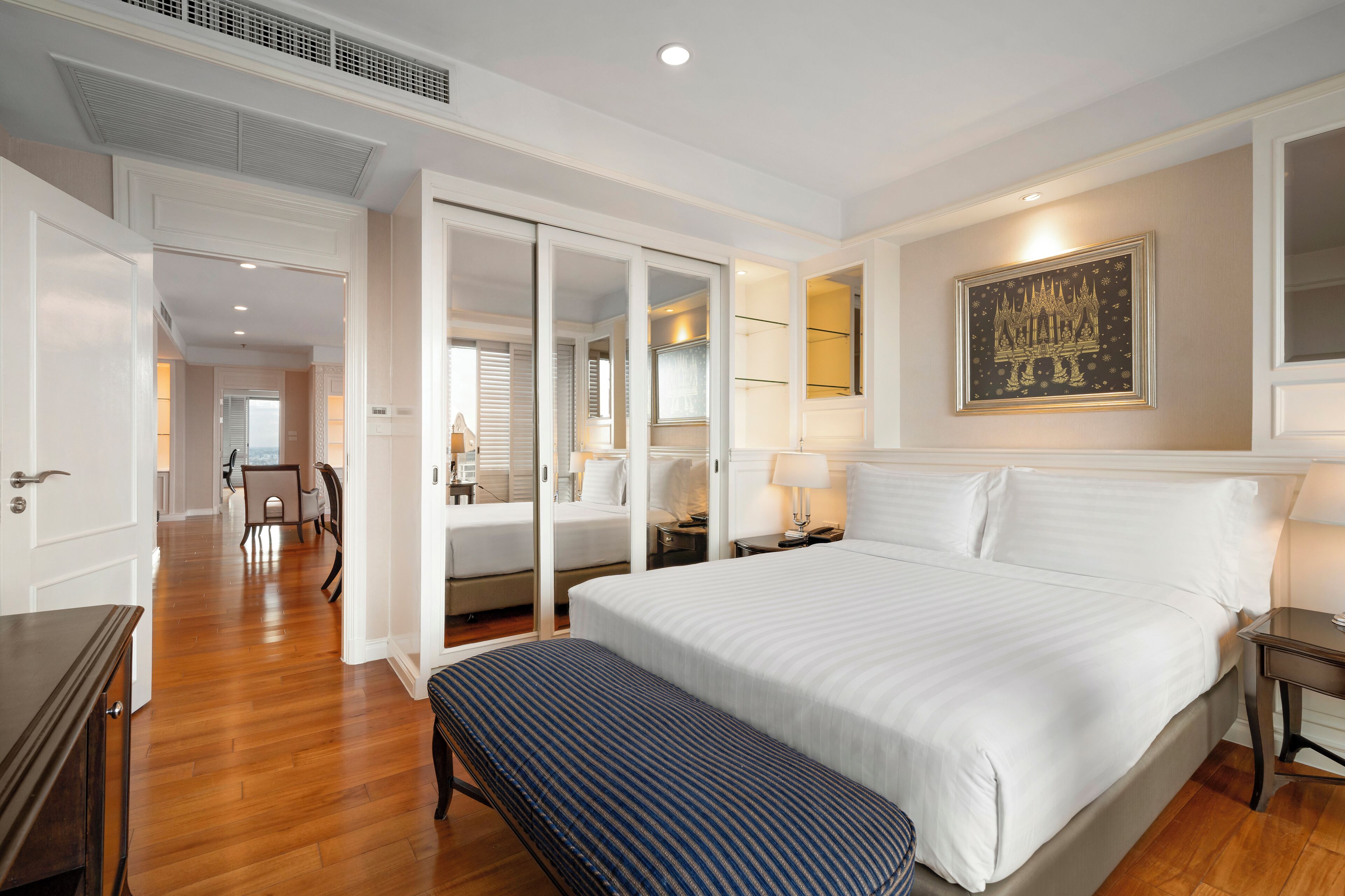 Une chambre d’hôtel avec un grand lit, une banquette, un bureau et une chaise. On y trouve une armoire avec un miroir et un tableau accroché au mur.