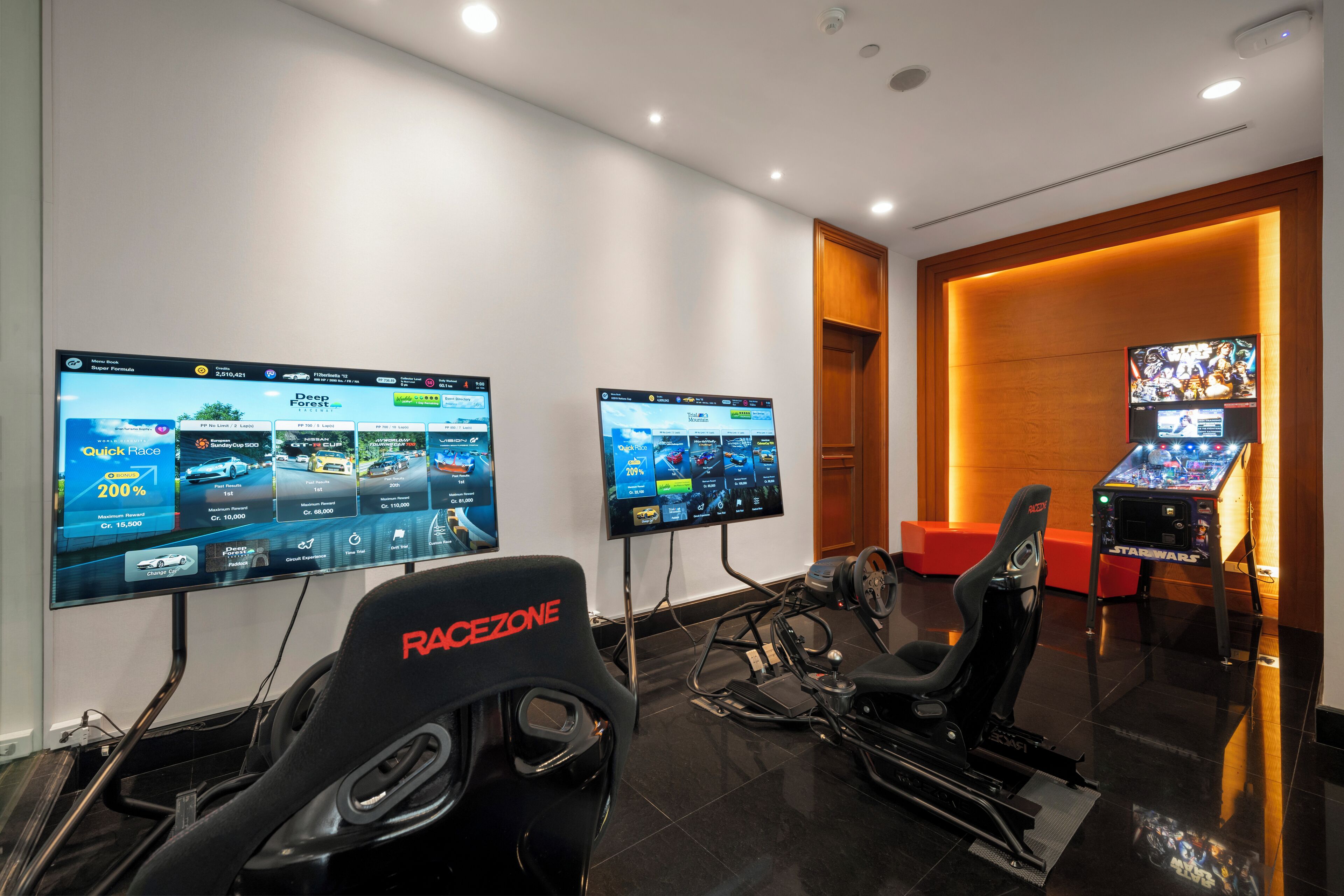 A gaming room with two large monitors displaying racing games, a racing seat, a racing simulator, and a Star Wars arcade game.
