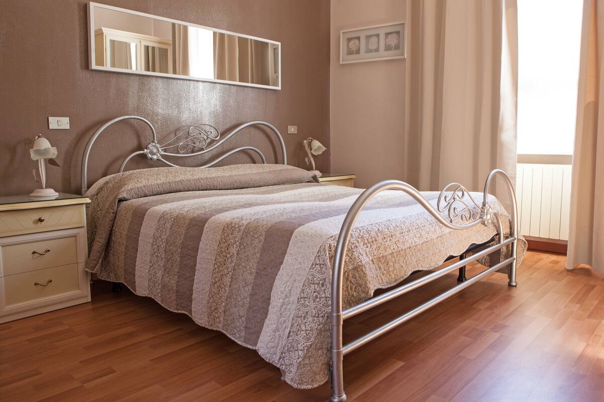 Un dormitorio con estructura de cama metálica, una mesita de noche con lámpara, un aparador con cajones y un espejo grande en la pared.
