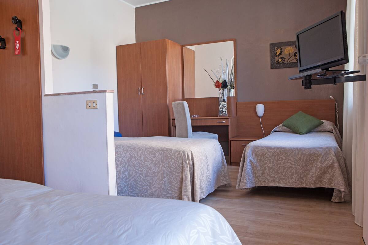 Una habitación de hotel con dos camas individuales, un escritorio con silla, un televisor montado en la pared y un armario.