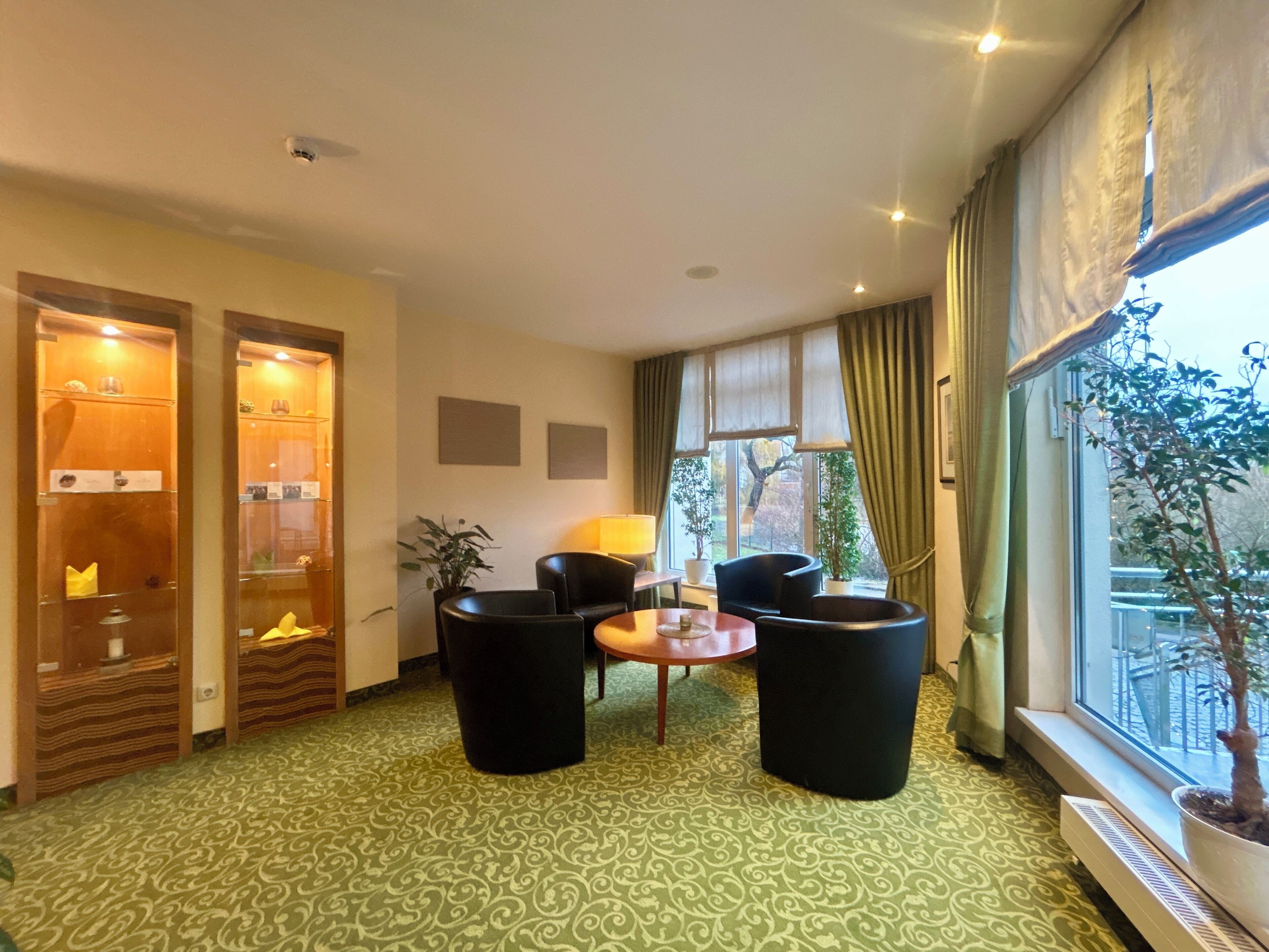 A hotel room with a green patterned carpet, two black armchairs, a small table, a window with curtains, and a potted plant by the window.