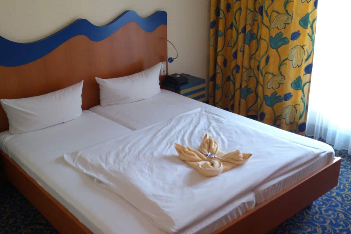 A hotel room with a bed, pillows, a nightstand, a lamp, and curtains.