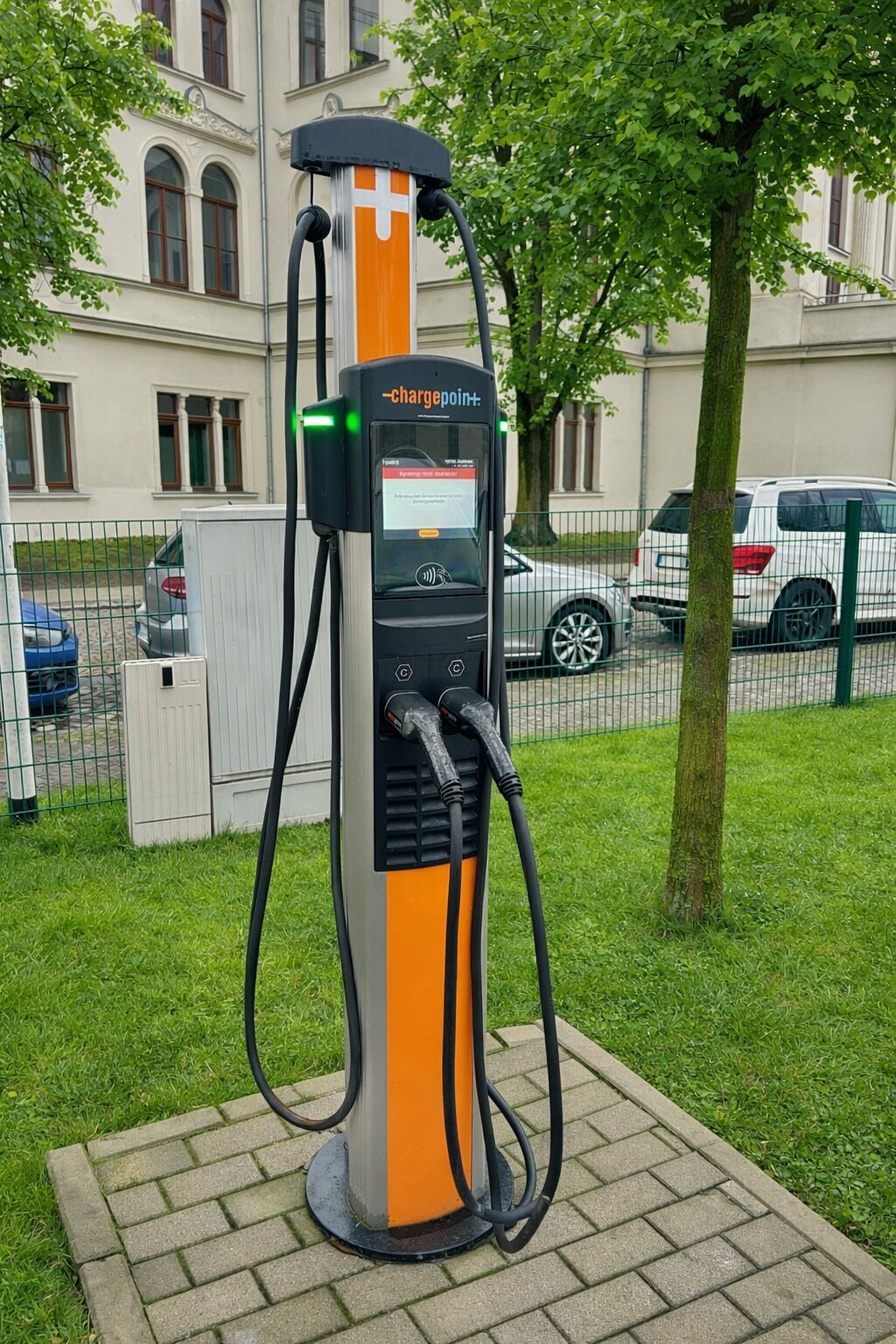 Una estación de carga pública para vehículos eléctricos, con pantalla digital, ubicada en un área verde con un edificio y coches estacionados al fondo.