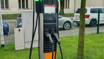 Una estación de carga pública para vehículos eléctricos, con pantalla digital, ubicada en un área verde con un edificio y coches estacionados al fondo.