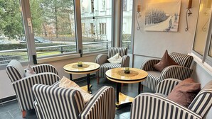 Een moderne zithoek met gestreept beklede stoelen en ronde tafels, grote ramen met uitzicht op een gebouw en bomen, en een schilderij aan de muur.