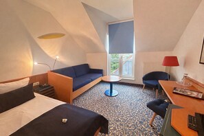 A hotel room with a bed, a sofa, a small table, a chair, and a window with a view of a building. - Hotel Am Kurpark Bad Salzelmen (Schönebeck)