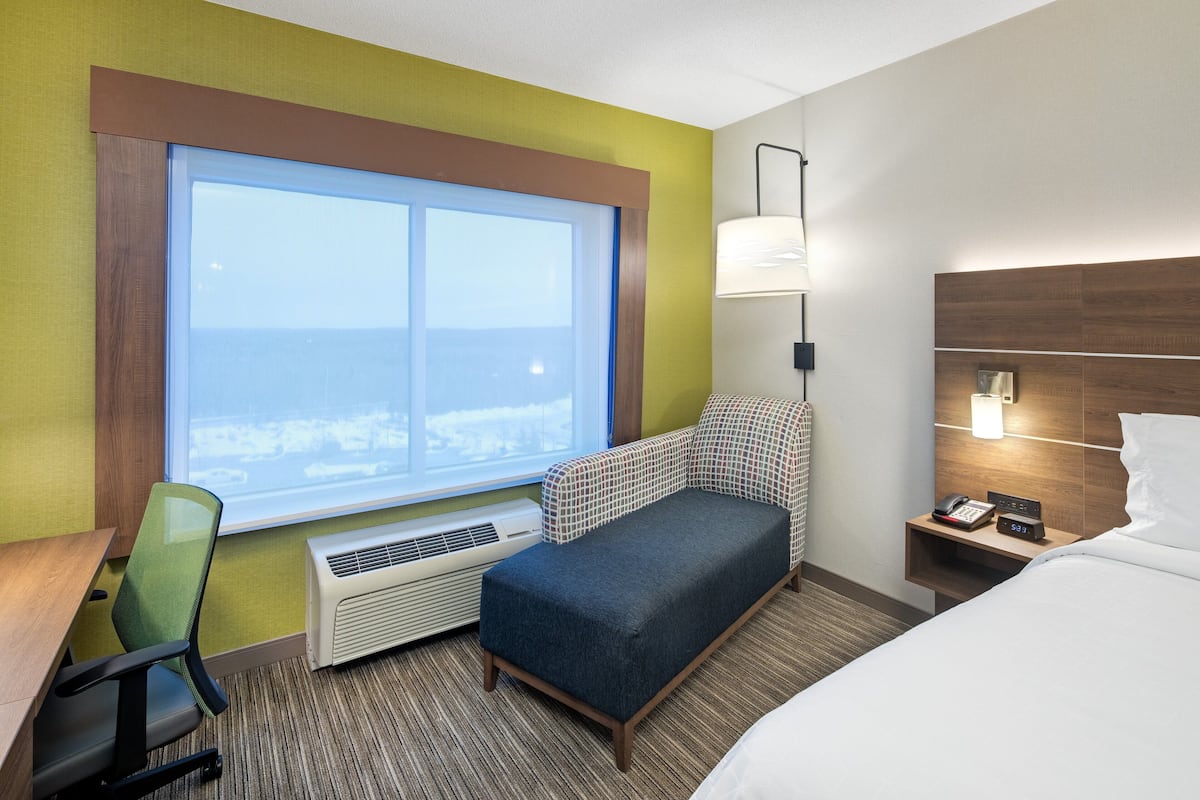 A hotel room with a bed, a desk with a chair, a small sofa, a nightstand with a lamp, and a window with a view.