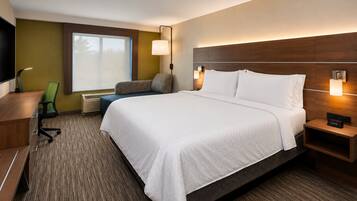 A hotel room with a large bed, a desk, a chair, a TV, and a window with blinds.