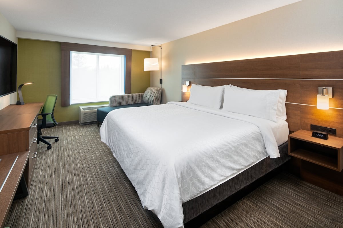 A hotel room with a large bed, a desk with a chair, a small table, and a nightstand with a lamp.