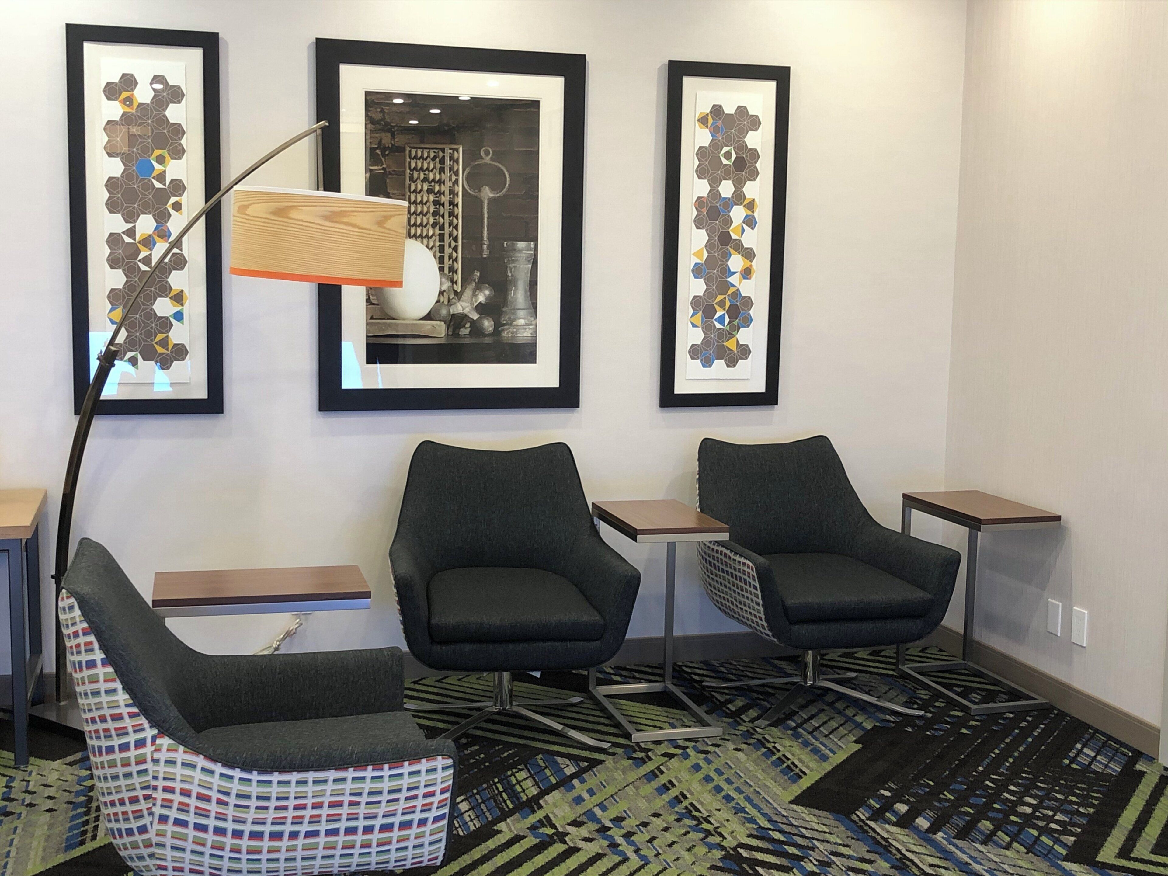 A hotel room with two armchairs, a small table, a floor lamp, and three framed artworks on the wall.