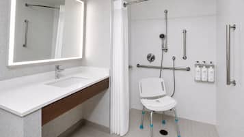 A modern bathroom with a large mirror, a sink, a shower with grab bars, and a white chair.