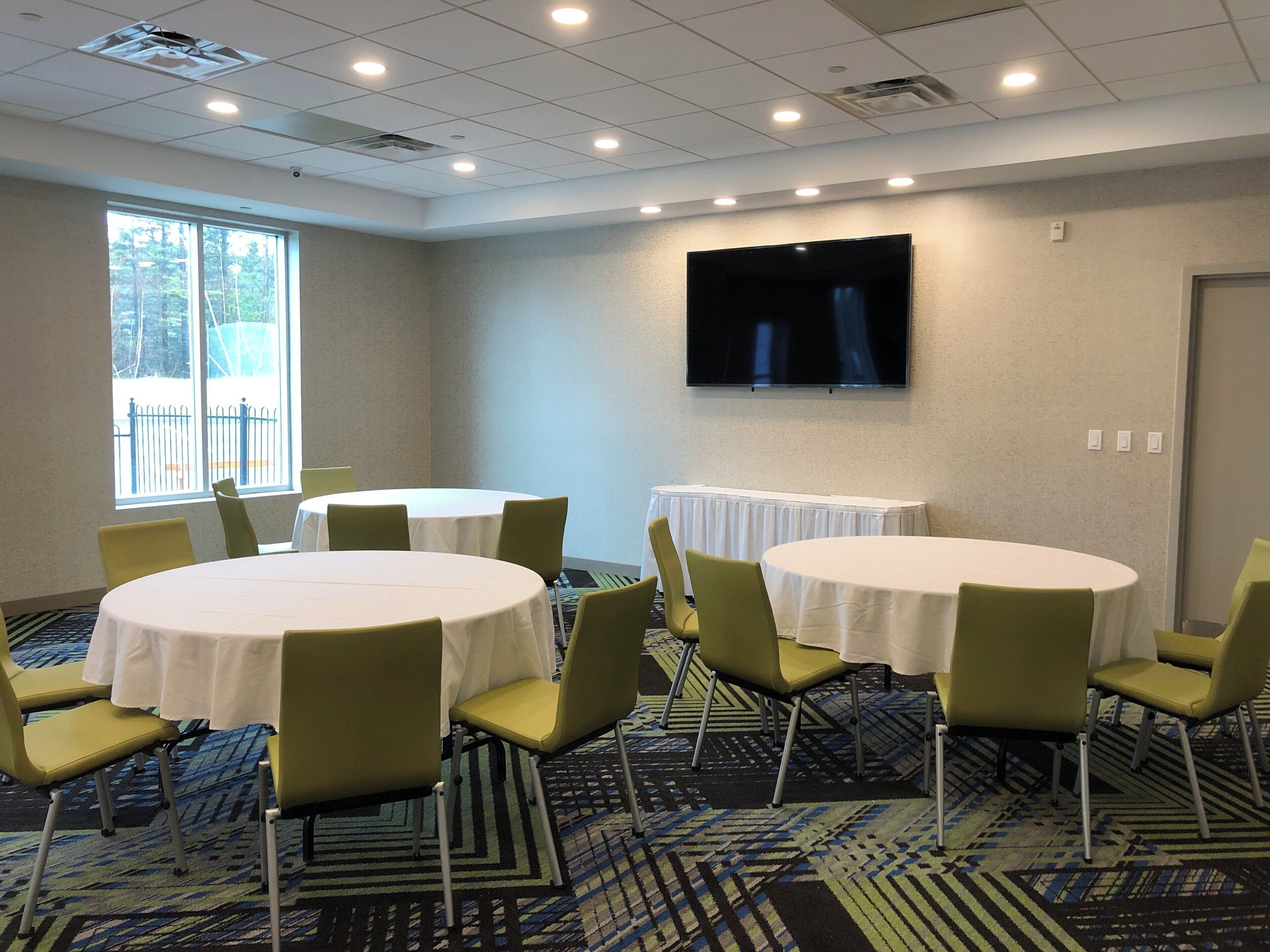 A conference room with round tables, green chairs, a flat-screen TV, and a large window with a view of trees.