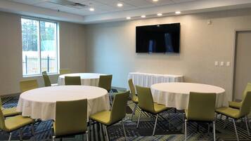 A conference room with round tables, green chairs, a flat-screen TV, and a large window with a view of trees.