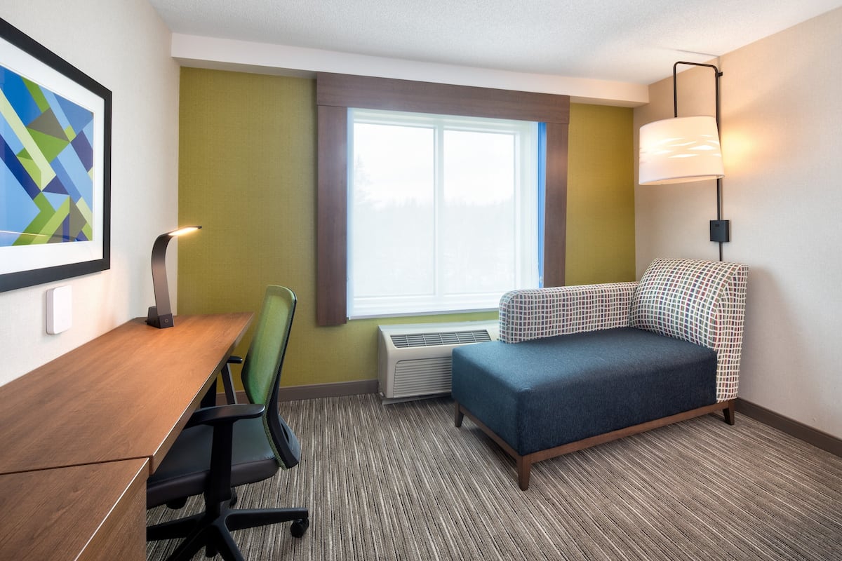 A hotel room with a desk, chair, sofa, window, lamp, and air conditioning unit.