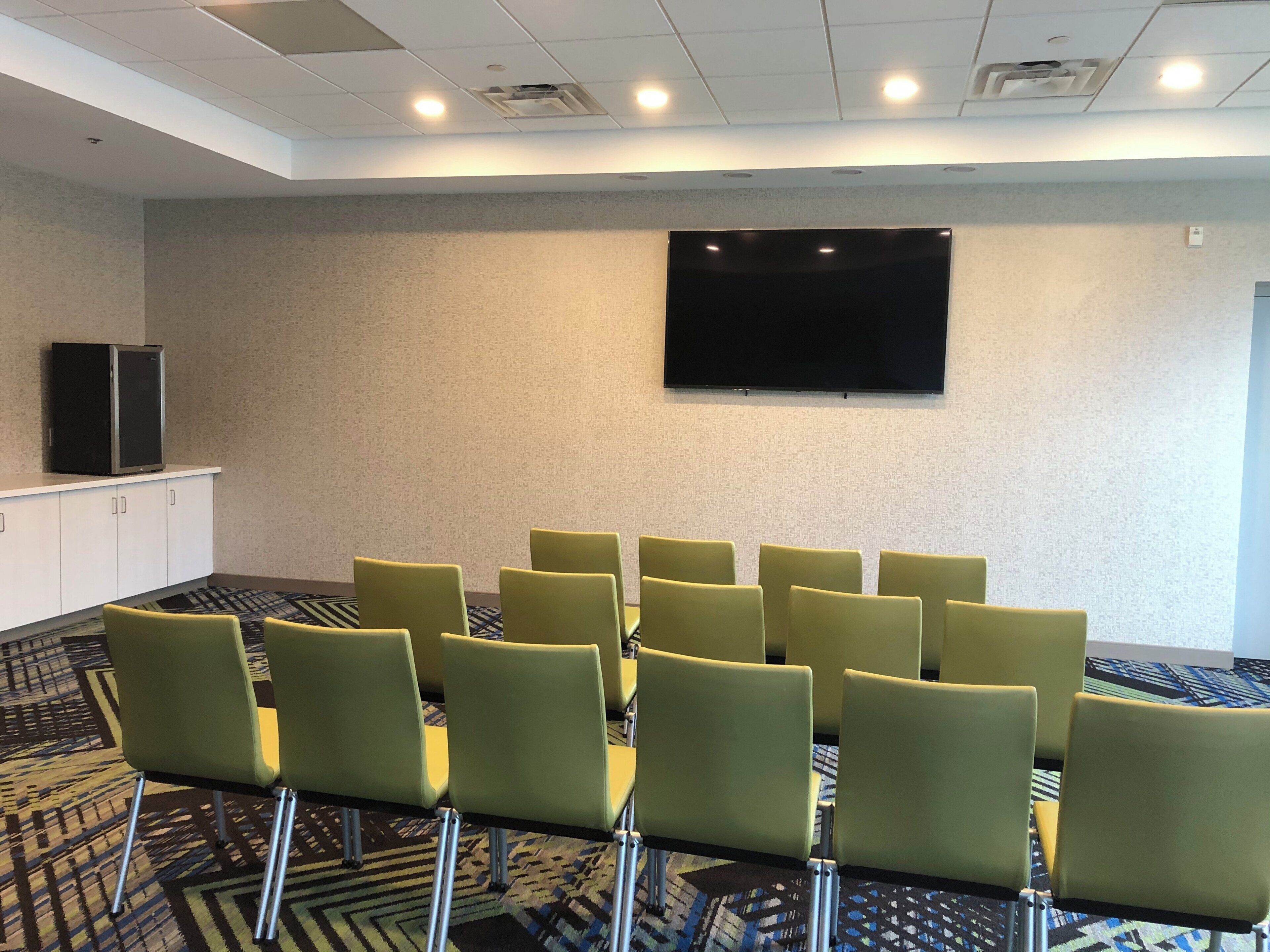 A conference room with a flat-screen TV mounted on the wall, a row of green chairs, and a patterned carpet.