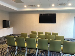 A conference room with a flat-screen TV mounted on the wall, a row of green chairs, and a patterned carpet.