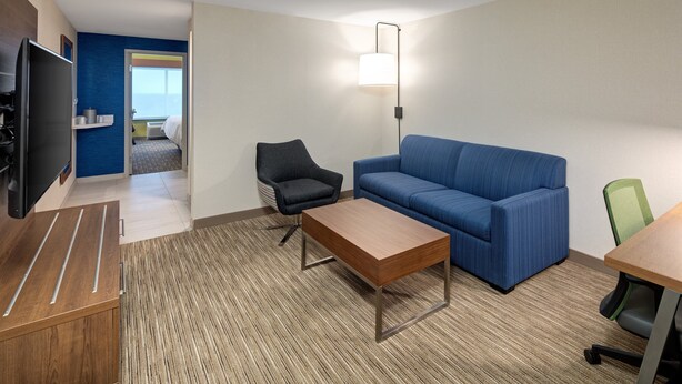 A hotel room with a blue sofa, a wooden coffee table, a black office chair, a green office chair, a flat-screen TV, and a lamp. - Holiday Inn Express & Suites Halifax Airport by IHG (Goffs)
