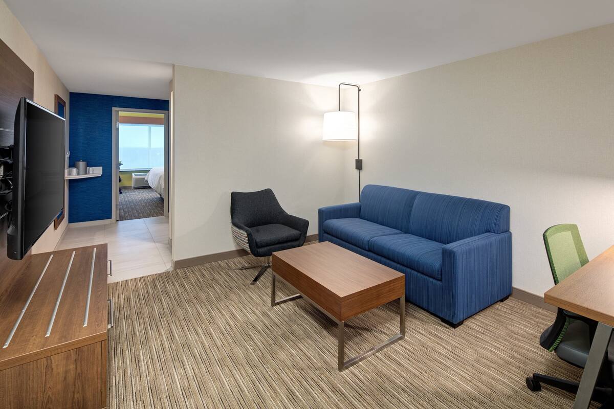 A hotel room with a blue sofa, a wooden coffee table, a black office chair, a green office chair, a flat-screen TV, and a lamp.
