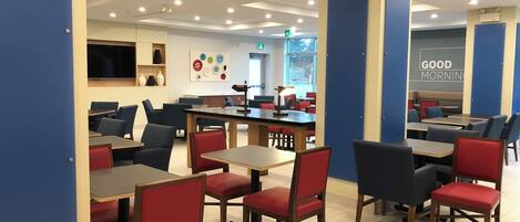 A modern cafe with multiple tables and chairs, a flat-screen TV mounted on the wall, and a 'Good Morning' sign on the wall.