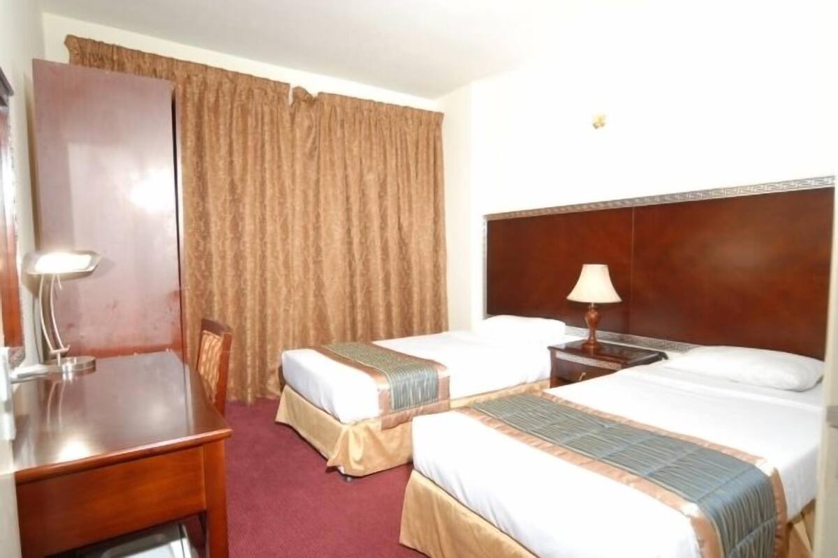 Une chambre d’hôtel avec deux lits, un bureau, une chaise, une lampe et une grande fenêtre avec des rideaux.