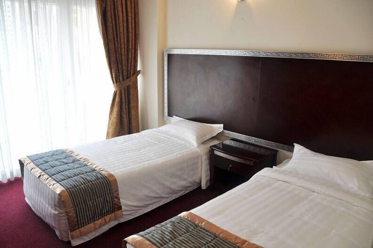 Une chambre d’hôtel avec deux lits, une tête de lit sombre, une table de chevet et une fenêtre avec des rideaux.
