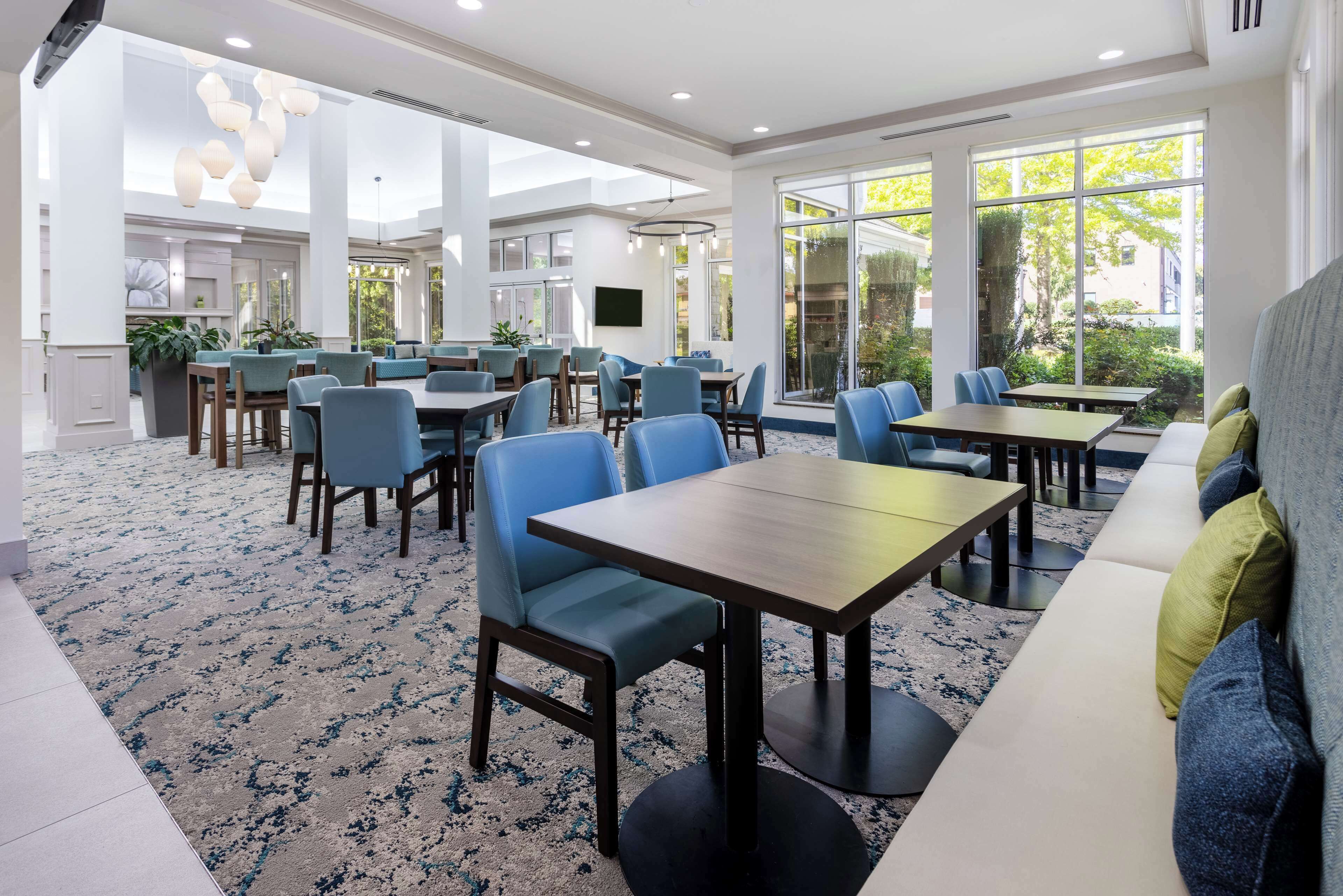 A spacious hotel dining area with multiple tables and chairs, large windows providing natural light, and a modern interior design.