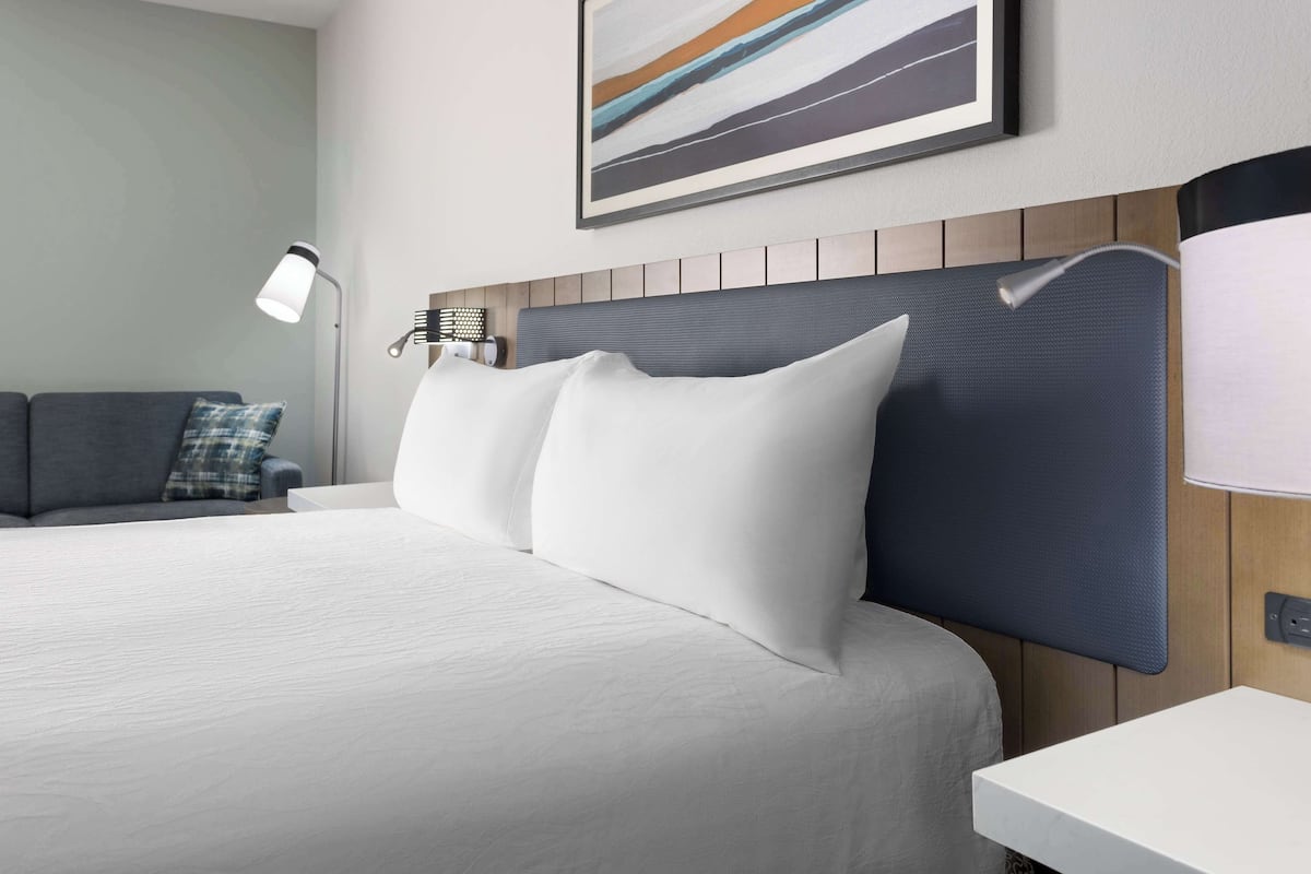 Een netjes opgemaakt bed met witte lakens, een hoofdbord met een geribbelde panelen, een nachtkastje met een lamp, en een grijze bank met een ruitkussen.