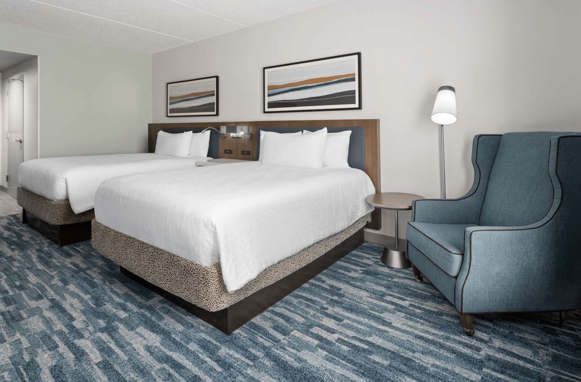 A hotel room with two beds, a blue armchair, a small round table, and two framed artworks on the wall.