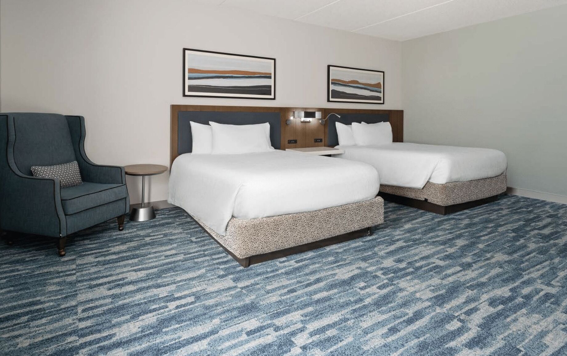 Een hotelkamer met twee bedden, een stoel, een kleine tafel en twee ingelijste foto’s aan de muur.