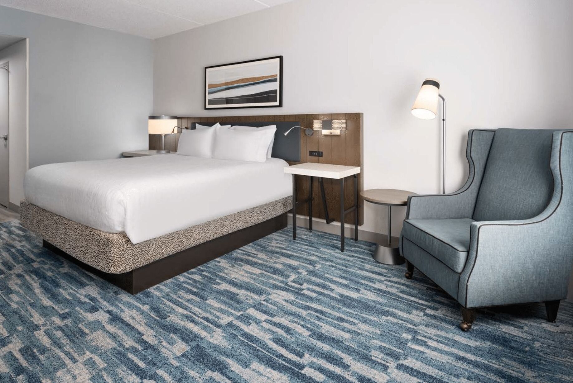 A hotel room with a large bed, a desk, a chair, and a lamp.