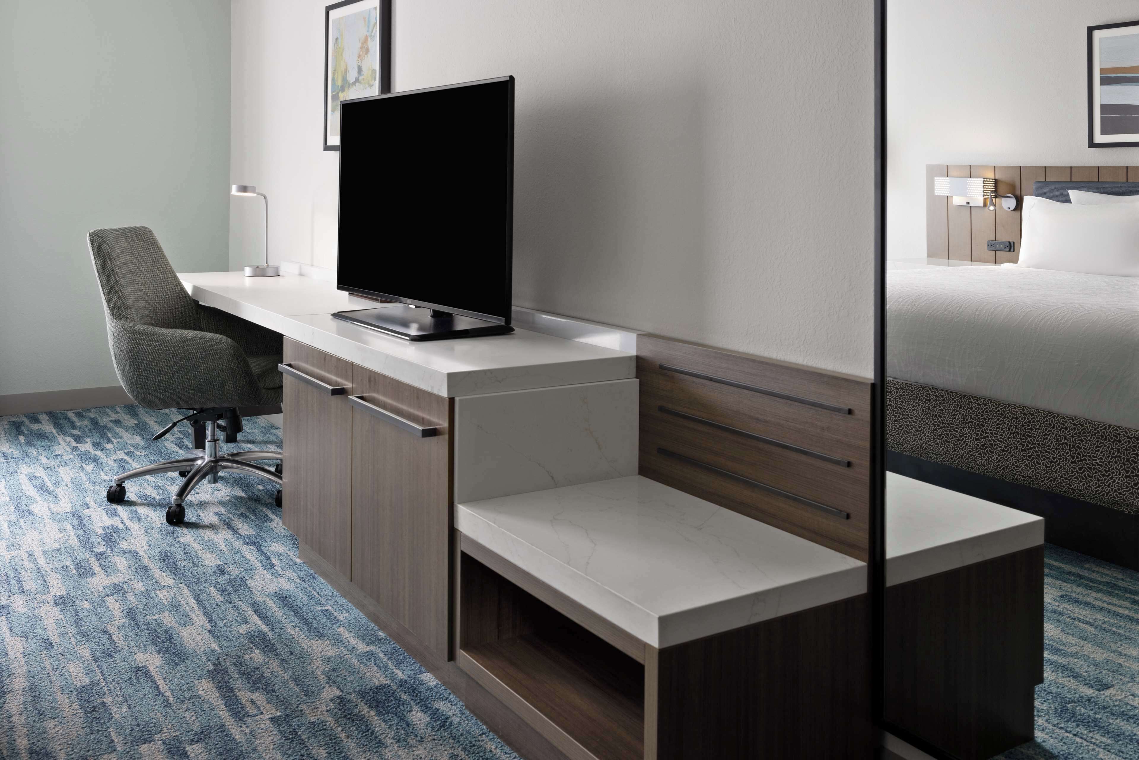 A hotel room with a desk, chair, TV, bed, and a nightstand with a lamp.