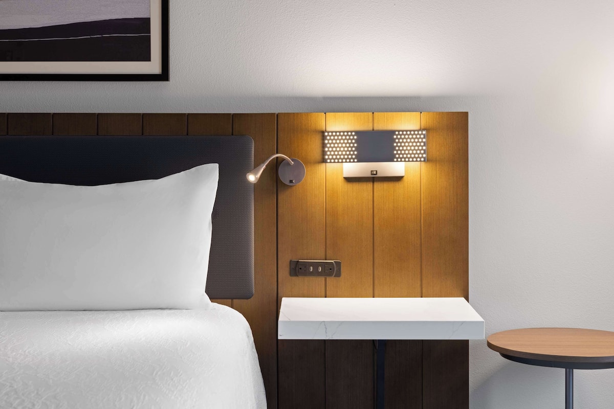 Hotelkamer met een bed, nachtkastje, een klein rond tafeltje en een wandlamp.