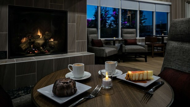 Un rincón comedor acogedor con chimenea, dos tazas de café, un postre y una vela sobre la mesa.