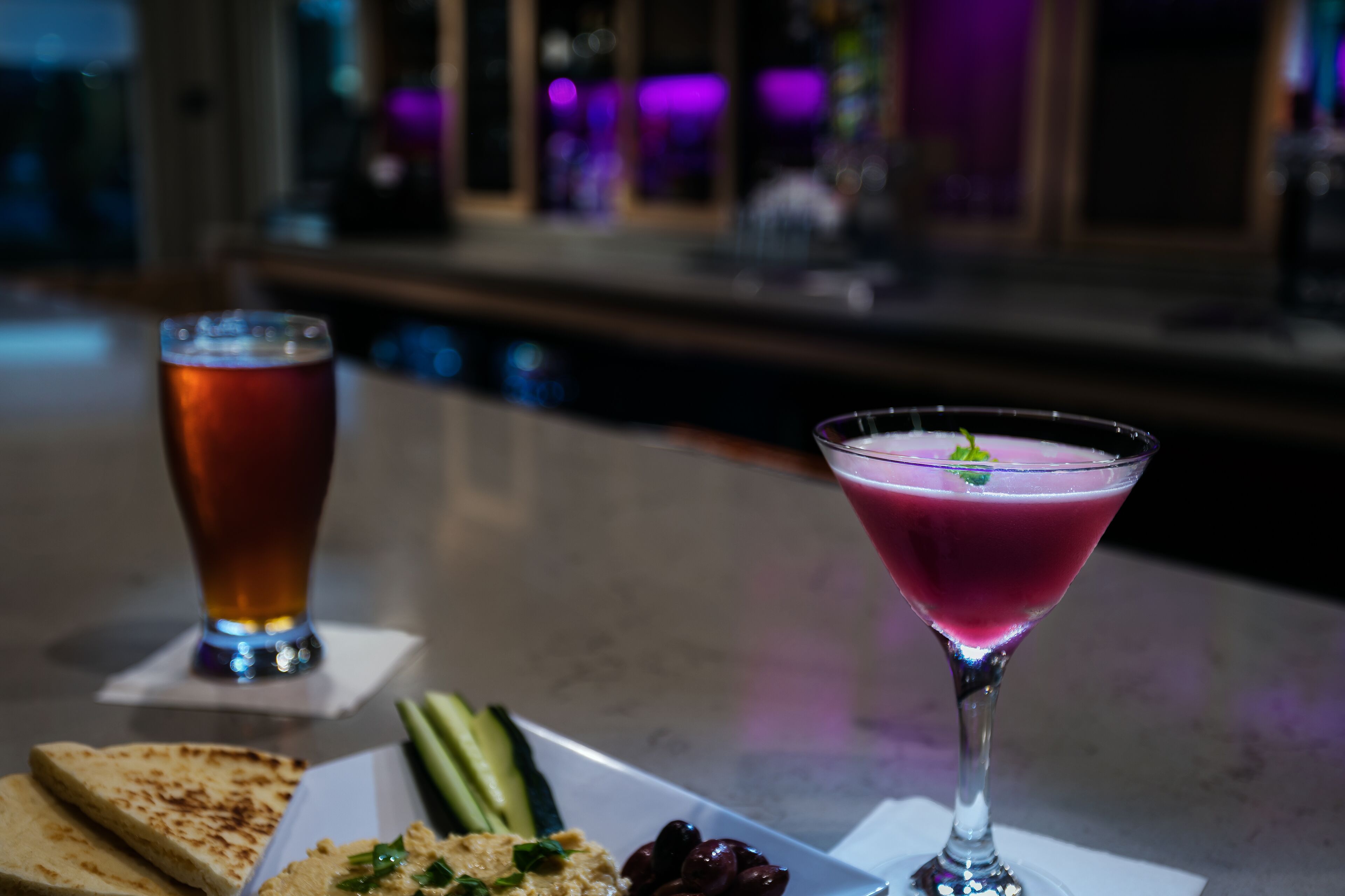 A bar setting with a martini glass and a beer glass on a table, accompanied by a plate of food including toast and vegetables.