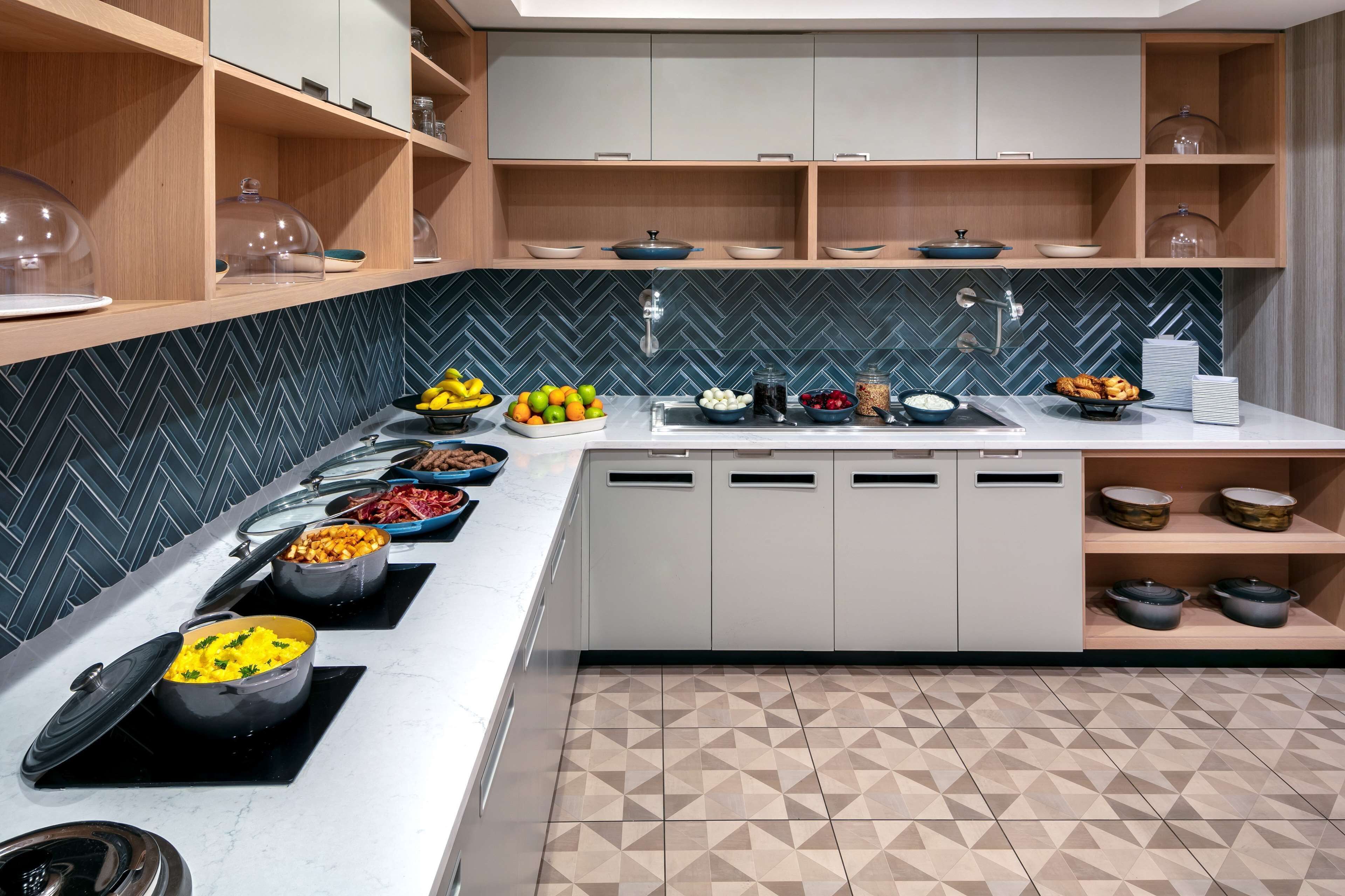 A modern kitchen with a herringbone tile backsplash, a long counter with various food items, and a built-in oven.