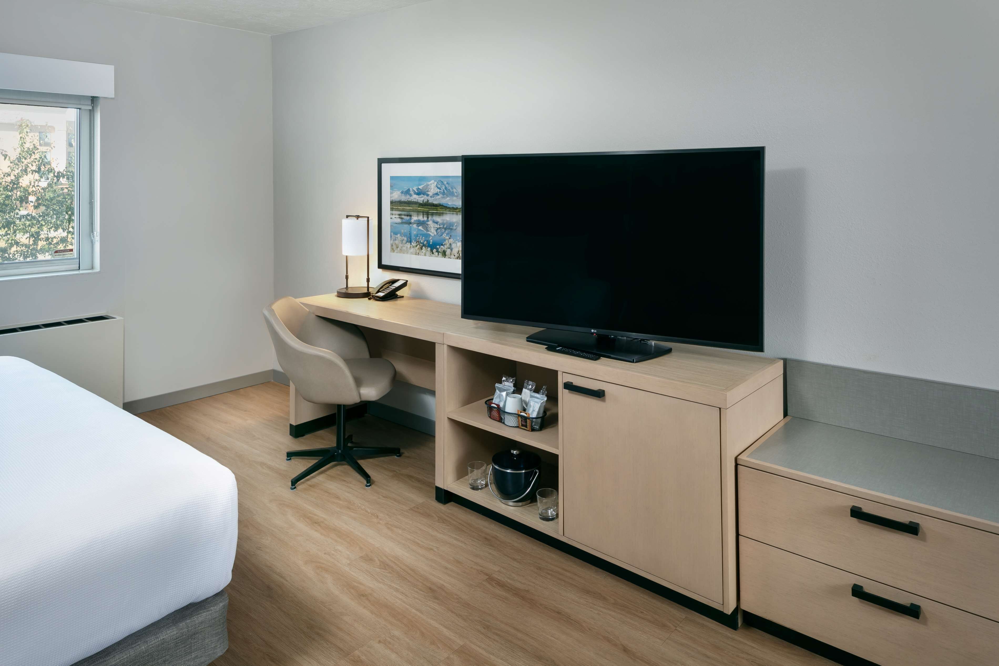 A hotel room with a bed, a desk with a flat-screen TV, a chair, a shelf with kitchen items, and a window with a view of trees.