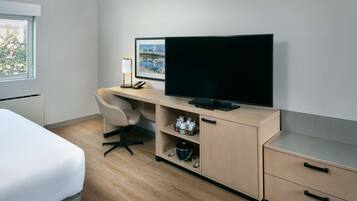 A hotel room with a bed, a desk with a flat-screen TV, a chair, a shelf with kitchen items, and a window with a view of trees.