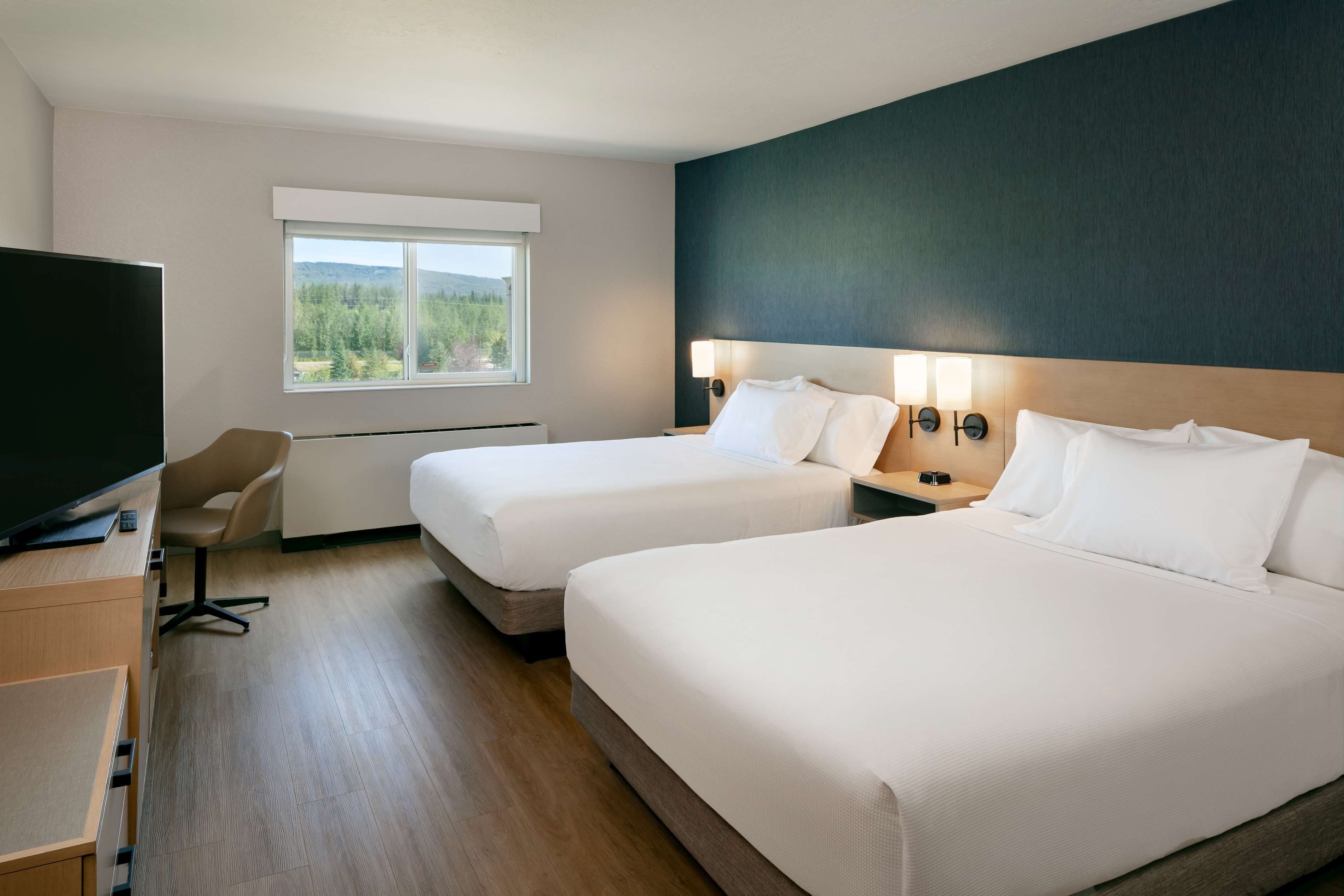 A hotel room with two beds, a desk with a TV, a chair, and a window with a view of trees and hills.